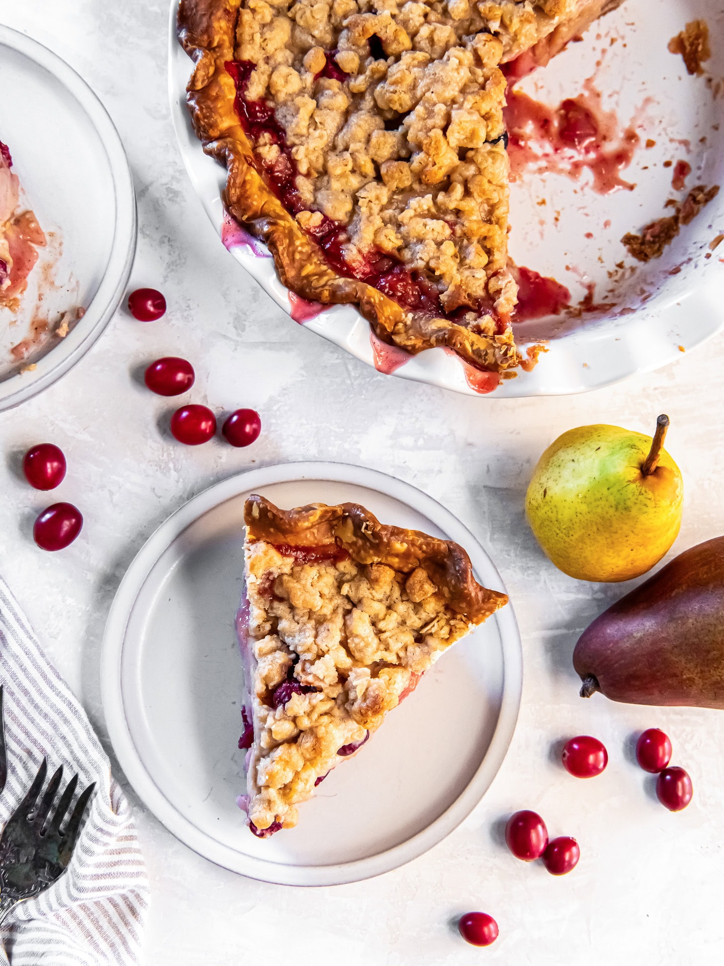 Pear pie with cranberries served out of a white pie plate onto white plates near fresh pears and cranberries.
