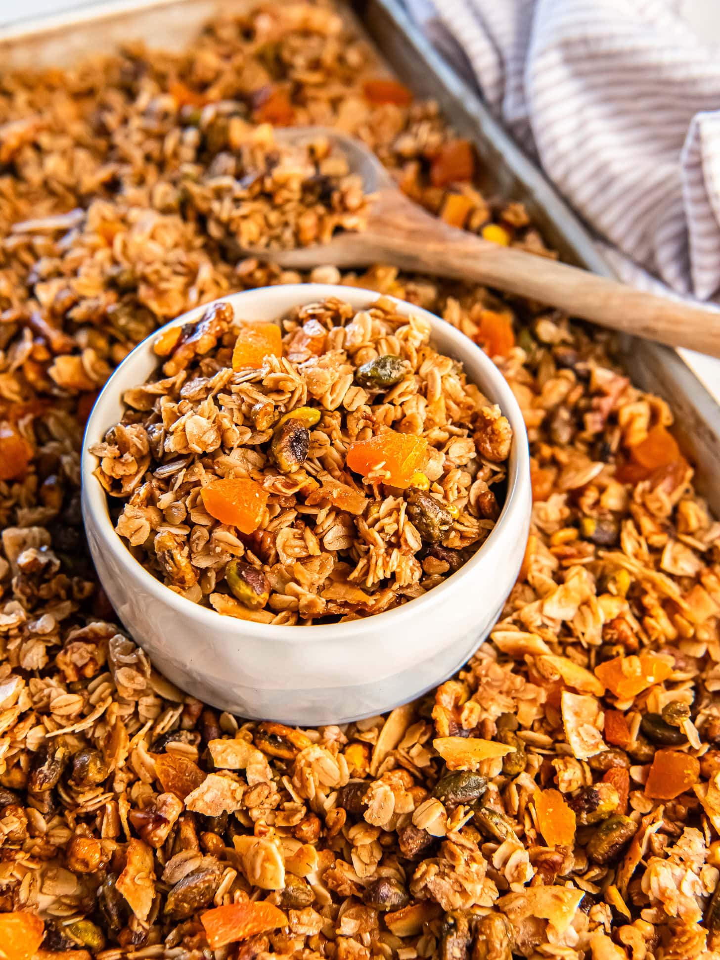 Apricot pistachio granola in a white bowl on a cookie sheet with more granola.