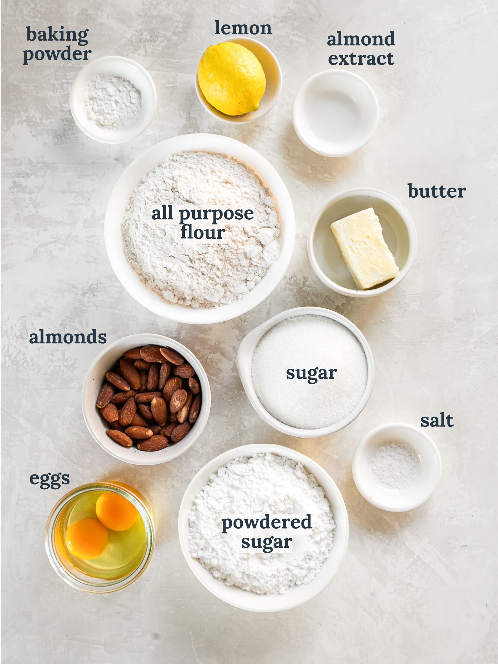 Small bowls with flour, almonds, sugar, eggs, butter and a lemon.