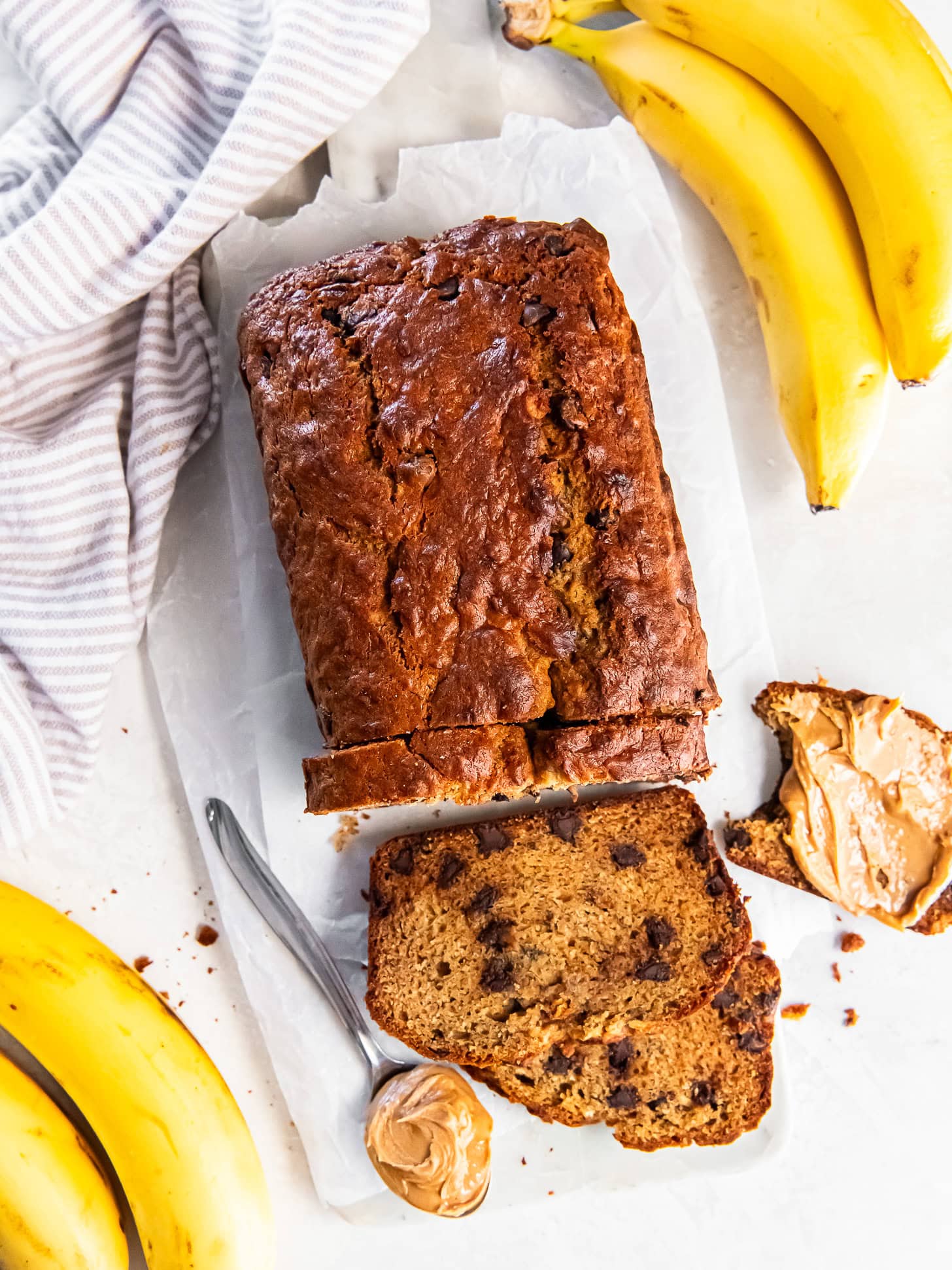 Loaf of sliced banana bread near fresh bananas and peanut butter.