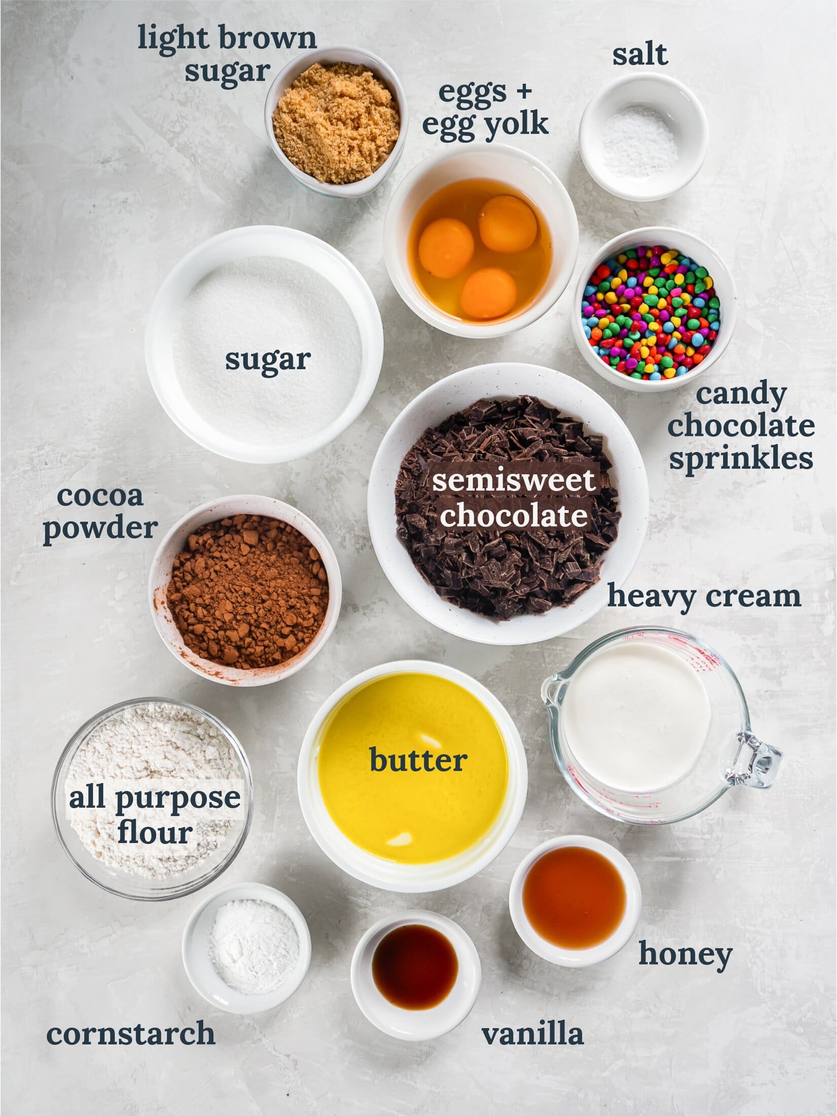 Small bowls with chocolate chips, sugar, cocoa powder, eggs, candy sprinkles, flour and cream.