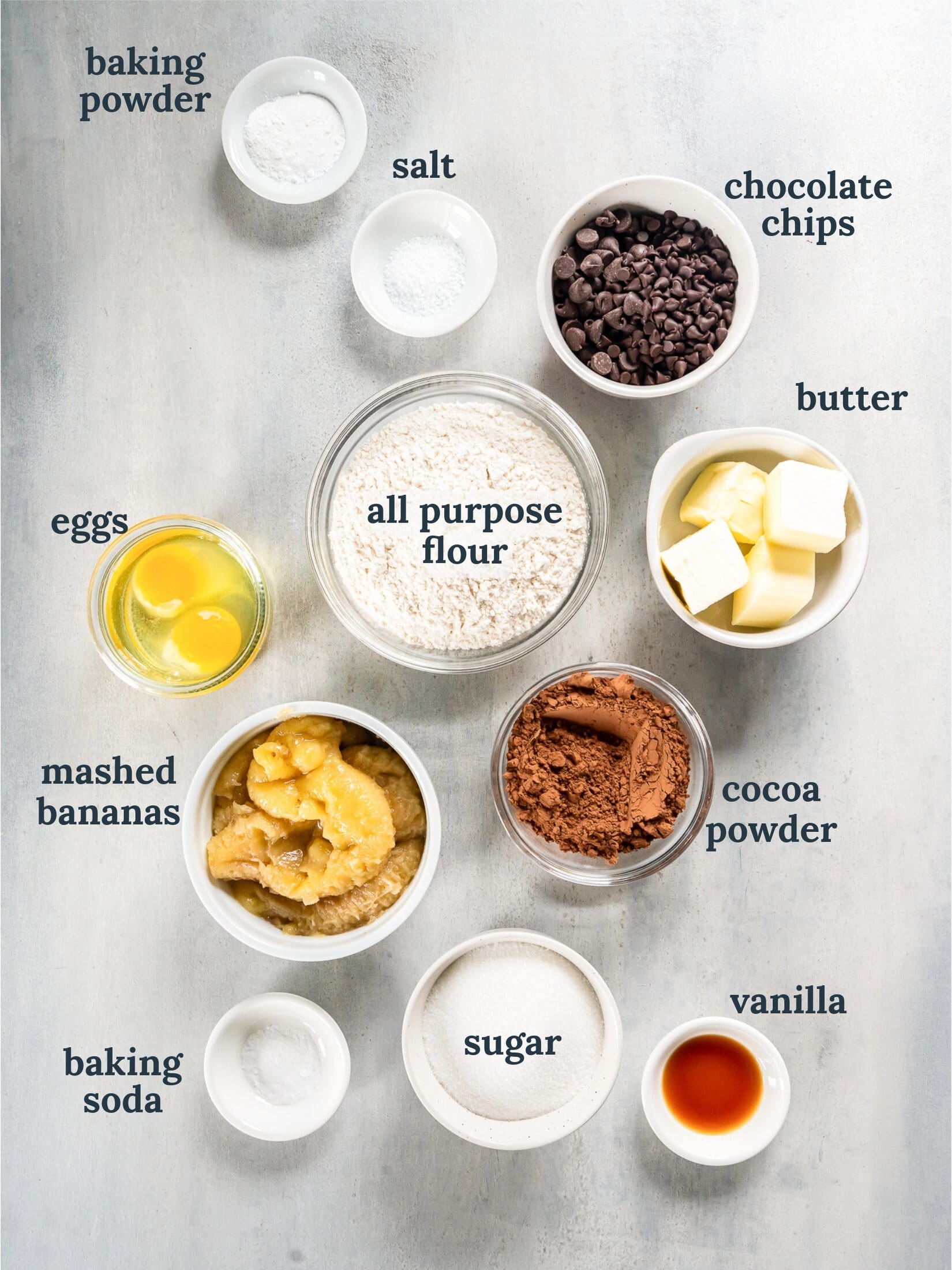 Small bowls with cocoa powder, flour, sugar, butter, eggs, mashed banana and chocolate chips.