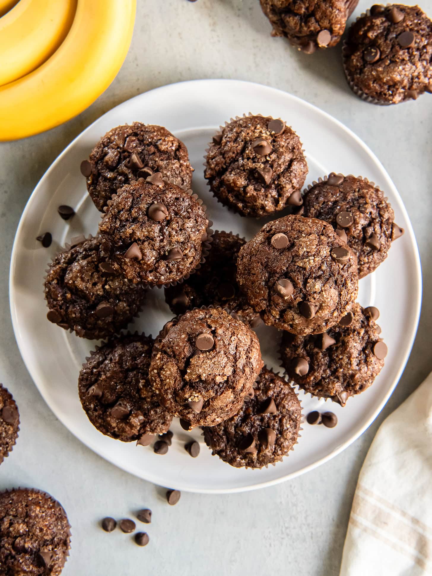 Double chocolate banana muffins stacked on a white plate near a bunch of bananas.
